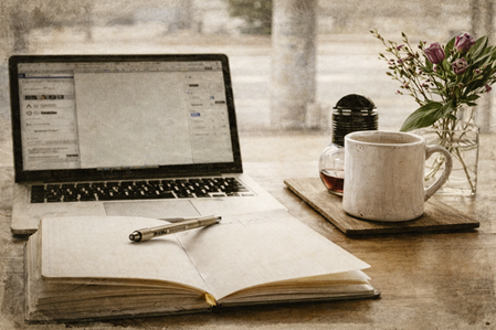 Laptop, aufgeschlagenes Notizbuch und eine Tasse Tee am Fenster